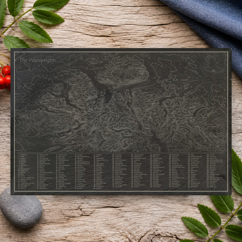 The Wainwrights natural slate placemat on a pale oak table with blue linen and rowan leaves: a contour map of the Lake District with all 214 fells numbered and listed in a key
