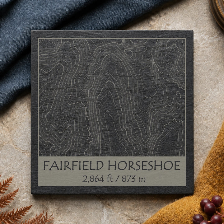 Fairfield Horseshoe topographic slate coaster on warm limestone with blue textile, bracken and green leaves
