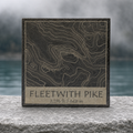 Rendered Fleetwith Pike contour-map design on a square natural slate coaster on a weathered stone ledge with a misty blue-green background