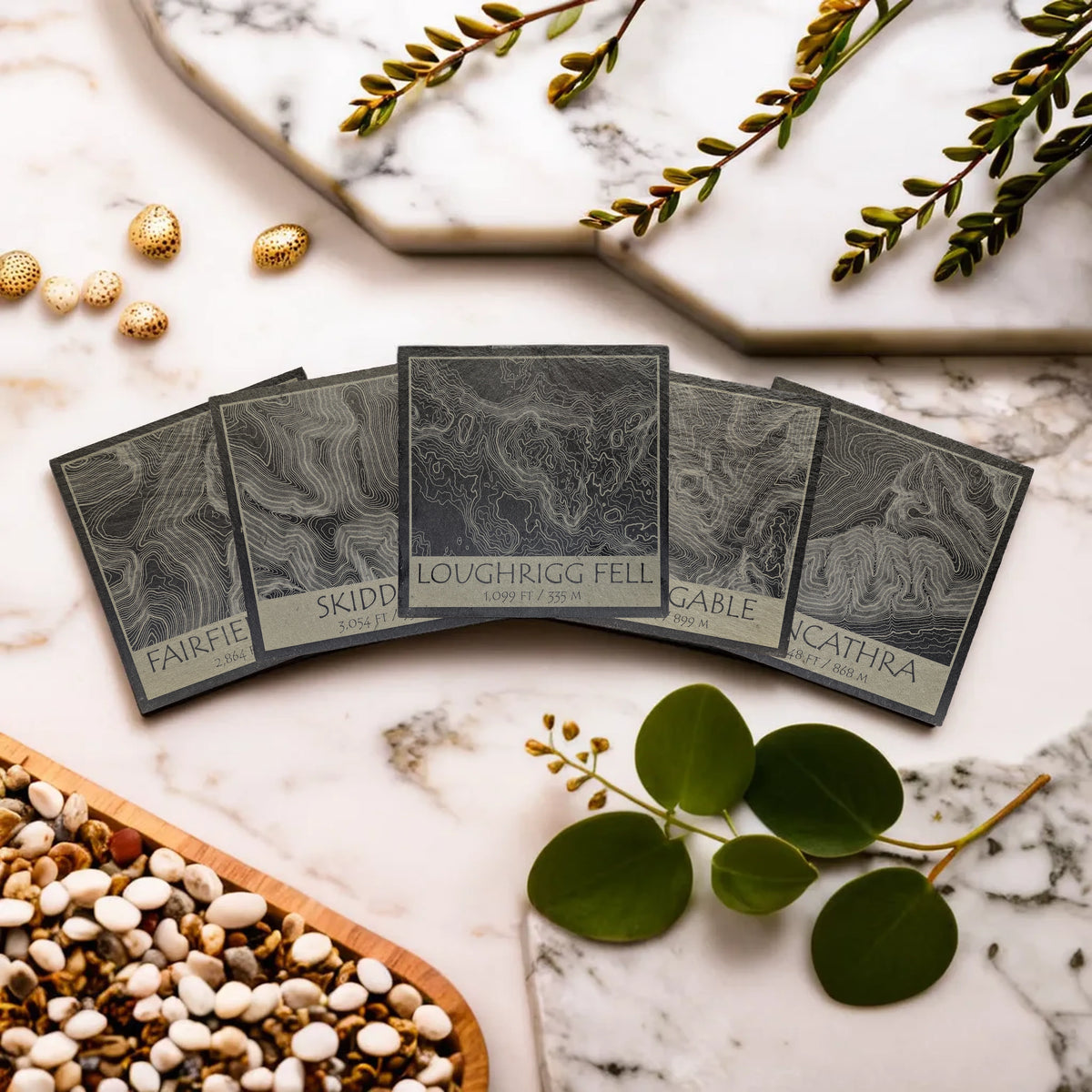 Five Lake District slate coaster designs arranged on a pale stone surface